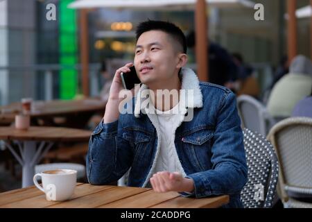 Nahaufnahme eines hübschen asiatischen jungen Mannes, der im Straßencafé sitzt und einen Anruf macht. Hintergrund verwischen Stockfoto