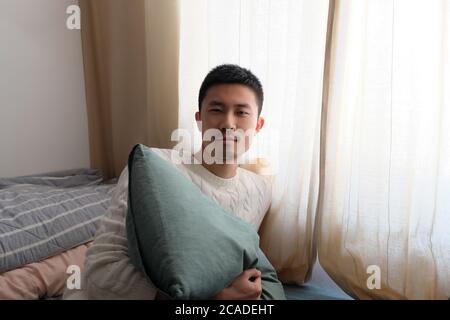 Porträt eines asiatischen jungen Mannes, der Kissen im Sitzen hält Bett im Schlafzimmer Stockfoto