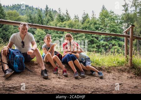 Portrait von lächelnden Eltern und ihren beiden niedlichen Kindern, die auf einem Pfad sitzen und eine Pause von ihrer Familienwanderung machen Stockfoto