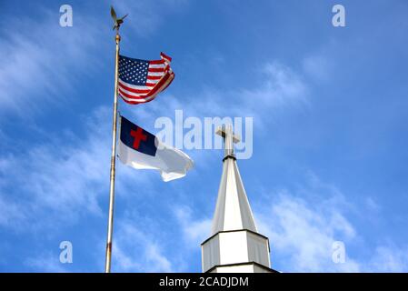 Amerikanische und christliche Flagge fliegen neben einem Kirchturm mit einem Kreuz-Symbol auf der Oberseite. Blauer Himmel und weiße, flüsterige Wolken füllen den Hintergrund. Stockfoto