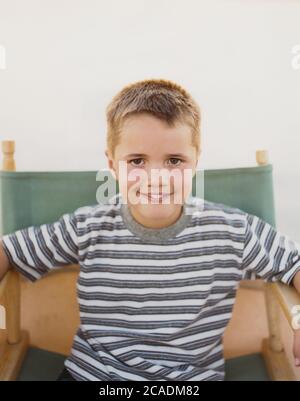 Ein selbstbewusster Junge, der in einem klappbaren Regiestuhl sitzt und ein toothy Lächeln lächelt. Stockfoto