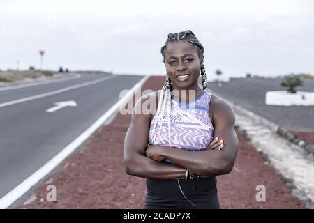 Selbstbewusste afroamerikanische Athletin, die die Arme kreuzt und für die Kamera lächelt, während sie während der Laufpause am Straßenrand steht Stockfoto