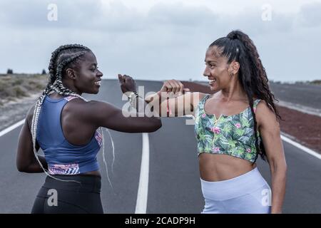 Glückliche multiethnische Freundinnen in Sportswear lächeln und stoßen Ellbogen, während sie sich beim Laufen Training auf der Straße grüßen Stockfoto