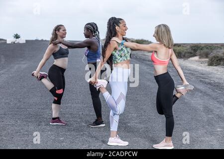 Seitenansicht fröhlicher multirassischer Freundinnen in Sportkleidung, die sich gegenseitig anlehnen und lächeln, während sie gebeugte Beine vor dem Laufen strecken Stockfoto