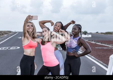 Gruppe von positiven, vielfältigen Frauen in Sportbekleidung, die Bizeps und Lächeln beim Selfie beim Outdoor-Lauftraining demonstrieren Stockfoto