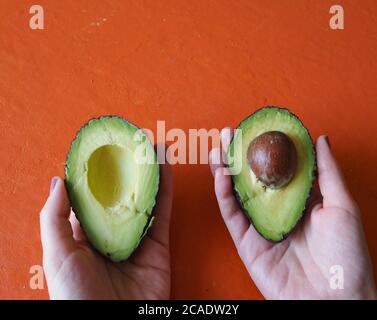 Weibliche Hände mit jeweils einer Hälfte Avocado Auf einem orangefarbenen Hintergrund Stockfoto