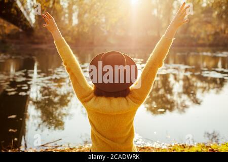 Reisende entspannen am Herbstfluss bei Sonnenuntergang. Glückliche Frau Tourist sitzt auf Bank, die Arme zu heben Gefühl frei Stockfoto