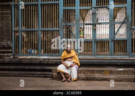 Kumbakonam, Tamil Nadu, Indien - Februar 2020: Ein älterer indischer Tempelpriester in traditioneller Kleidung sitzt neben einer großen Grilltür insid Stockfoto