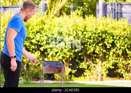 Schöner Athlet männlichen Kochen Grill im Freien. Brutaler bärtiger Mann stand und bereitete Fleisch auf dem Hof zu. Rückansicht. Speicherplatz kopieren. Stockfoto