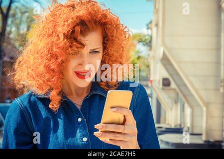 Was. Unglücklich frustriert verärgerte Frau mit roten lockigen Haaren Blick auf Handy, SMS lesen oder so etwas, stehen außerhalb der Stadt auf dem Hintergrund. Negat Stockfoto
