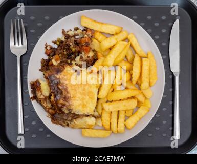 Nahaufnahme POV über dem Kopf Aufnahme von einem Messer und Gabel neben einem Teller auf einem Tablett, von heißen Cottage Pie mit Gemüse, begleitet von Chips / Pommes. Stockfoto