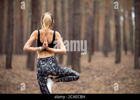 Schöne Frau, die im Wald steht und Yoga macht. Übung und Meditation Konzept. Befolgung zahlen oder Hand heben Konzept. Kiefernholz im Sommerthema. Rückansicht. Stockfoto