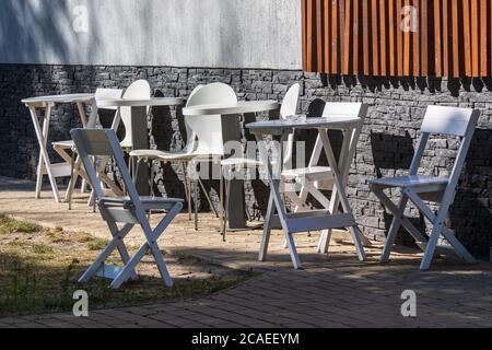 Tische und Stühle aus weißem Holz stehen an einem sonnigen Sommertag ohne Besucher an einer grauen Steinmauer. Der Terrassenbereich des Coffeeshops. Ein gemütliches Café. Ein Ort t Stockfoto