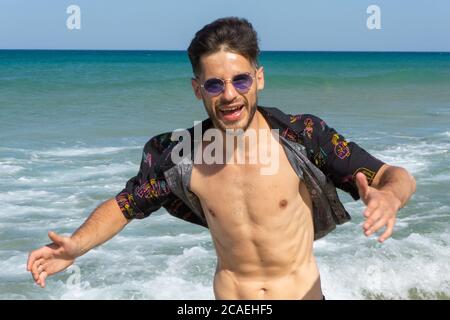 Glücklicher junger Mann in Sonnenbrille mit Spaß am Strand Mit dem Meer auf seinem Hintergrund Stockfoto