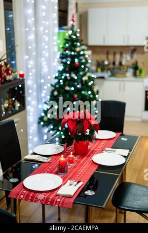 Ein gemütliches Zimmer mit Tischgestelltem für das neue Jahr vor dem Hintergrund eines festlich geschmückten Weihnachtsbaums mit Girlanden und einer weißen Küche. Stockfoto