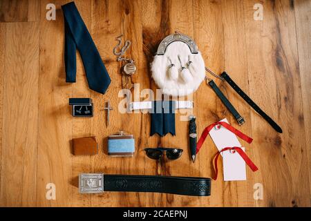 Scotsman Sporran's Accessoires, Taschenuhr an einer Kette, Krawatte, Manschettenknöpfe, Kreuz, Gürtel, Messer, Kolben, Gläser, Brieftasche auf einer glatten Holzoberfläche. Stockfoto