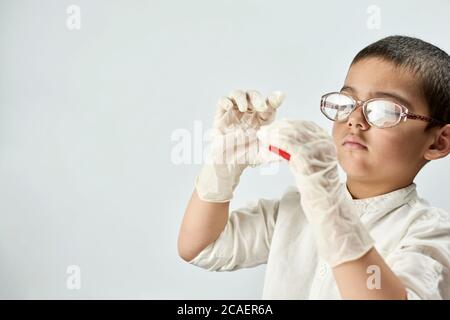 Ein Porträt eines niedlichen Jungen in Schutzhandschuhen und Brille mit einem Stück rotem Plastilin in den Händen vor dem weißen Hintergrund Stockfoto