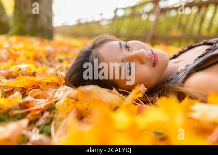 Junge schöne asiatische Frau, die nach oben schaut, während sie sich auf gelben Herbstblättern legt Stockfoto