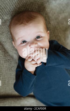 Neugeborenes Baby saugt Hände in den Mund, schönes Baby mit blauen Augen liegt auf grauen Wolldecken, Zahnfleisch jucken, Baby beißend Stockfoto