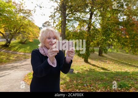 Ältere glückliche Frau lächelnd, während Sie mit dem Handy auf dem Weg des ruhigen und entspannenden Naturparks fotografieren Stockfoto