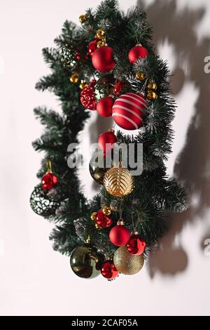 Grüner Neujahrskranz und Tannenzweige hängen an der weißen Wand, geschmückt mit Weihnachtsspielzeug, roten und goldenen Kugeln hängen, Glaskugeln hängen an Chr. Stockfoto