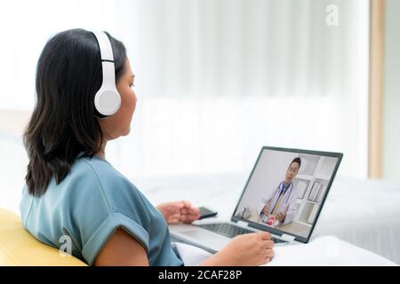 Rückansicht der asiatischen Frau macht Videoanruf mit ihrem Arzt mit ihrem Gefühl krank auf Laptop im Schlafzimmer für Online-Gesundheitswesen digitale Technologie-Service Stockfoto