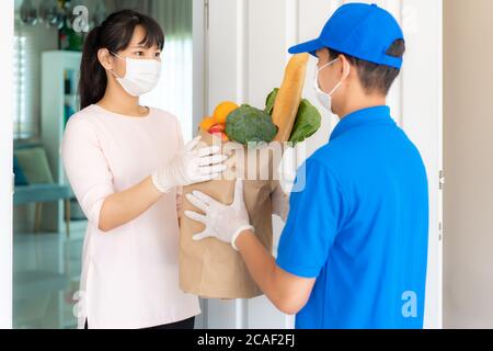 Asiatische Frau Kunden tragen Gesichtsmaske und Handschuh erhalten Lebensmittel Beutel mit Lebensmitteln, Obst, Gemüse und Getränke von der Lieferung Mann vor dem Haus d Stockfoto