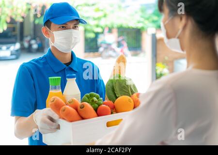 Asiatische Frau Kunde trägt Gesichtsmaske und Handschuh erhalten Lebensmittel-Box mit Lebensmitteln, Obst, Gemüse und trinken von Lieferung Mann vor dem Haus d Stockfoto