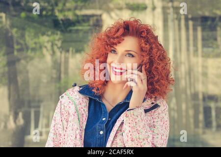 Image der jungen schönen fröhlichen Curly rothaarige Frau, die draußen im Park in der Nähe einer Bushaltestelle im Gespräch per Handy lächelnd. Stockfoto