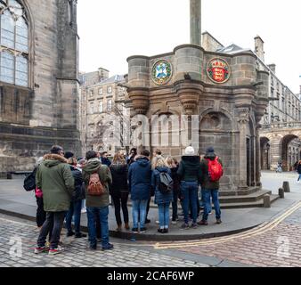 Eine Reisegruppe, die ihrem Fremdenführer vor dem Mercat Cross neben der St Giles Cathedral in der Royal Mile / High Street, Edinburgh, Schottland, Großbritannien zuhört Stockfoto