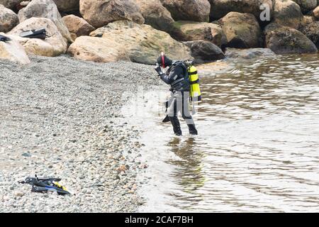 Ein Mann läuft aus dem kalten Atlantik mit Tauchausrüstung. Er hat einen gelben Sauerstofftank auf dem Rücken. Er entfernt seine Tauchermaske. Stockfoto