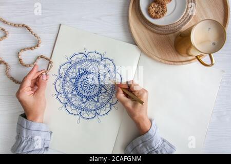 Weibliche Hände close up zeichnen Dekorative runde Blumenmandala. Hobby und Entspannung zu Hause. Eine Tasse Kaffee und Kekse auf einem Holztablett. Weißer Hintergrund. Stockfoto