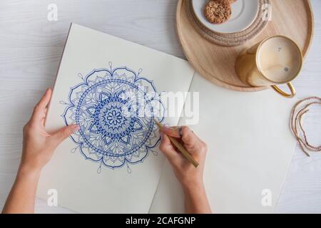 Weibliche Hände close up zeichnen Dekorative runde Blumenmandala. Hobby und Entspannung zu Hause. Eine Tasse Kaffee und Kekse auf einem Holztablett. Weißer Hintergrund. Stockfoto