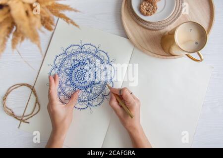 Weibliche Hände close up zeichnen Dekorative runde Blumenmandala. Hobby und Entspannung zu Hause. Eine Tasse Kaffee und Kekse auf einem Holztablett. Weißer Hintergrund. Stockfoto