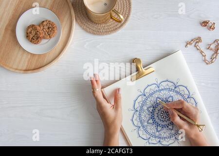 Weibliche Hände close up zeichnen Dekorative runde Blumenmandala. Hobby und Entspannung zu Hause. Eine Tasse Kaffee und Kekse auf einem Holztablett. Weißer Hintergrund. Stockfoto