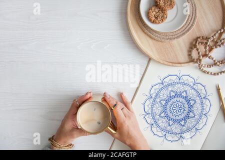 Weibliche Hände close up zeichnen Dekorative runde Blumenmandala. Hobby und Entspannung zu Hause. Eine Tasse Kaffee und Kekse auf einem Holztablett. Weißer Hintergrund. Stockfoto