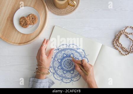 Weibliche Hände close up zeichnen Dekorative runde Blumenmandala. Hobby und Entspannung zu Hause. Eine Tasse Kaffee und Kekse auf einem Holztablett. Weißer Hintergrund. Stockfoto