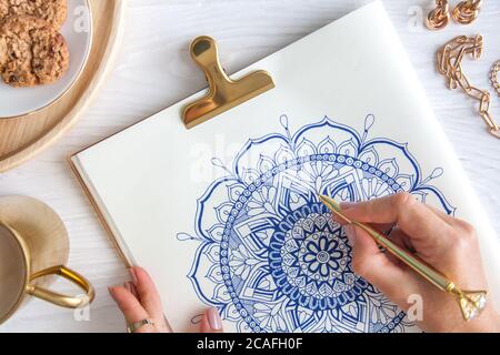 Weibliche Hände close up zeichnen Dekorative runde Blumenmandala. Hobby und Entspannung zu Hause. Eine Tasse Kaffee und Kekse auf einem Holztablett. Weißer Hintergrund. Stockfoto