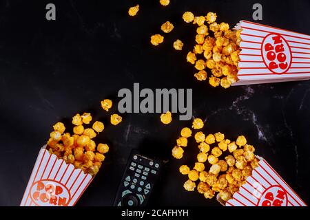 Popcorn in gestreiften Boxen verschüttet auf schwarzem Hintergrund mit Fernbedienung zum Fernsehen. Stockfoto