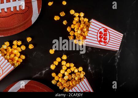 Gelbes Popcorn in gestreiften Schachteln auf schwarzem Hintergrund mit american Football Teller verschüttet. Sportspiel-Konzept ansehen. Stockfoto