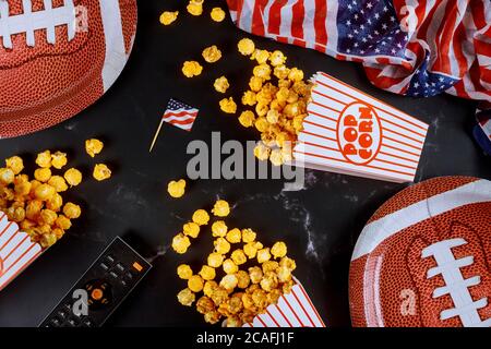 Gelbes Popcorn in gestreiften Schachteln auf schwarzem Hintergrund mit Fernbedienung und american Football-Teller verschüttet. Sportspiel-Konzept ansehen. Stockfoto