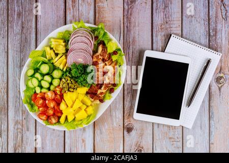 Notizbuch und Tablet mit gesalzenem Ernährungssalat auf Holztisch. Stockfoto