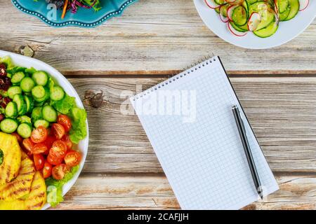 Notizbuch und Stift zum Schreiben von Rezepten mit gesunden Salaten auf Holztisch. Stockfoto