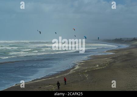 Kitesurfer reiten auf Wellen in Strandnähe, während die Leute im Vordergrund am Strand spazieren. Muriwai Beach, Westküste, Auckland, Neuseeland. Stockfoto