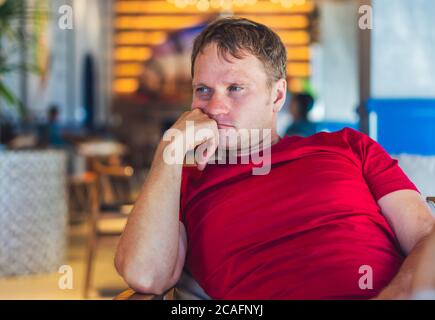 Serious blonde Mann im lässigen Stil roten T-Shirt sitzen im modernen Café, nervös jemand warten oder ändert, denken. Skin Small photoshop. Menschliche Beziehungen Stockfoto