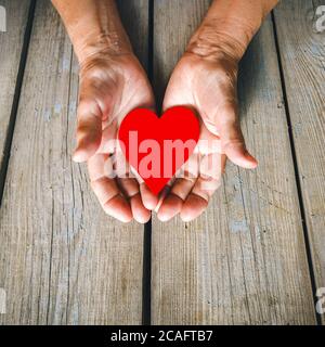 Ältere Frau Hände Nahaufnahme halten rote Herzen. Rustikale Holztisch Hintergrund. Liebe, Wärme, kümmern Konzept, Valentinstag, Muttertag, spenden, helfen. Stockfoto