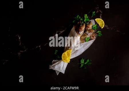 Ganzer frischer Fisch in Pergamentpapier auf schwarzem Hintergrund eingewickelt. Draufsicht. Stockfoto