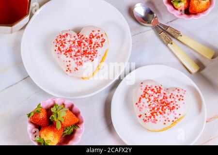 Zwei herzförmige Donuts mit weißer Glasur und rosa, roten Streuseln auf einer Platte mit Erdbeere und Löffel. Valentinstag-Konzept. Stockfoto