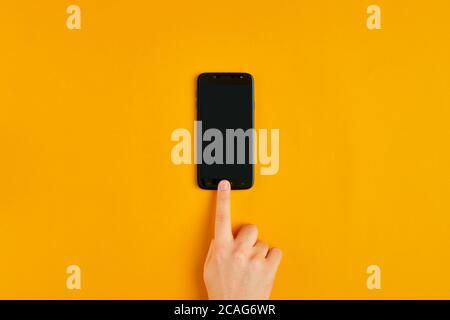 Männliche Hand drückt die Taste eines Smartphones mit leerem schwarzen Bildschirm auf gelbem Hintergrund. Stockfoto