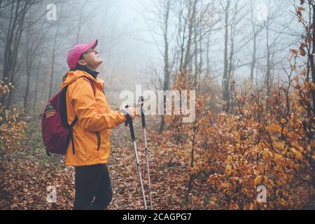 Dressed leuchtend orange Jacke junge weibliche Backpacker genießen die Natur. Sie läuft im Herbst Nebelwald mit Trekkingstöcken. Aktive Menschen und ein Stockfoto